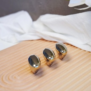 May include: Three polished, oval-shaped, silver-colored objects are arranged on a light-colored wooden tray. The objects reflect the surrounding environment. The tray has a curved design and is made of light-colored wood.