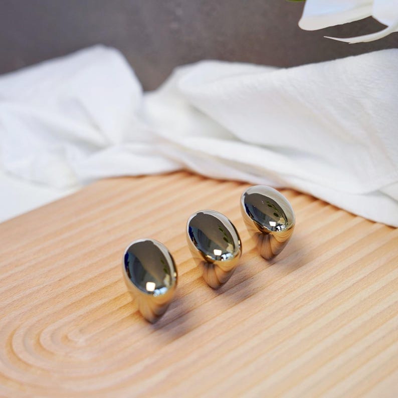 May include: Three polished, oval-shaped, silver-colored objects are arranged on a light-colored wooden tray. The objects reflect the surrounding environment. The tray has a curved design and is made of light-colored wood.