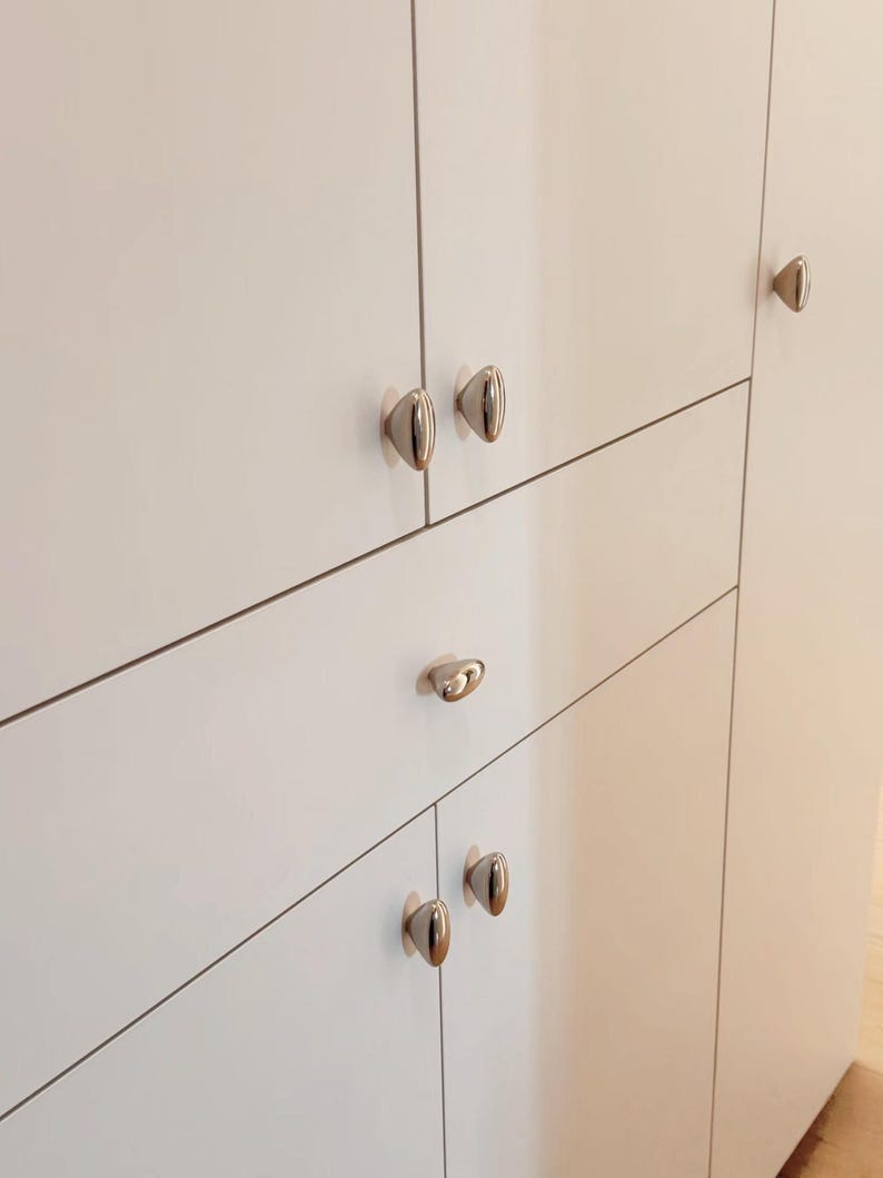 May include: Close-up of a white cabinet with several silver-colored oval cabinet knobs. The knobs are evenly spaced across the cabinet doors and drawers. The cabinet has a clean, modern design.