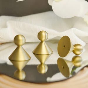 May include: Three gold-colored cabinet knobs with a geometric design. Each knob features a conical base topped with a spherical element. The knobs are arranged on a reflective surface, with their reflections visible.