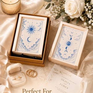 May include: Two rectangular wooden boxes with blue celestial illustrations of suns, moons, and clouds. One box is labeled Pilas, the other Caspian. A velvet ring box contains a gold ring, alongside two gold bands and a wedding invitation for Olivia and James.