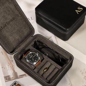 May include: A black rectangular jewelry storage box with a zippered closure and the gold initials AS on the lid. The interior is lined with dark gray fabric and contains a wristwatch with a brown leather strap, a ring, and black sunglasses.