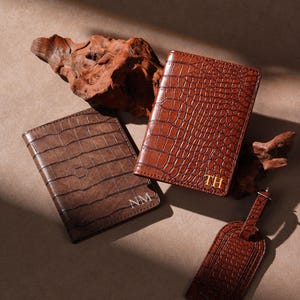Puede incluir: Se muestran tres artículos de cuero marrón con textura de cocodrilo. Se ven dos carteras y una etiqueta de equipaje. Las carteras están grabadas con iniciales, una con "NM" y la otra con "TH".