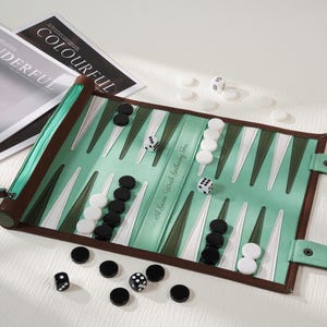 Puede incluir: Un tablero de backgammon enrollable en verde menta y marrón oscuro con fichas blancas y negras. El tablero presenta puntos triangulares en verde oscuro y blanco, dos dados blancos y el texto A Game Worth Gathering For impreso en él.