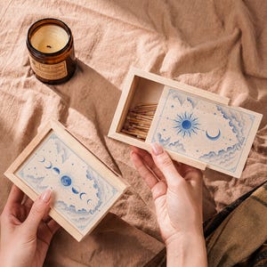 May include: Two rectangular light-colored wooden matchboxes with sliding lids. One box is open, revealing wooden matches inside. Both lids feature blue ink illustrations of celestial bodies, clouds, and stars, with the text Caspian and Silas.