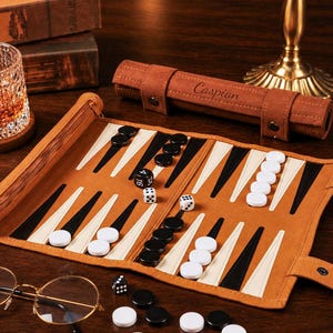 May include: A tan roll-up backgammon board featuring black and cream-colored triangular points. The set includes black and white circular checkers, two white dice, and one black doubling die. A matching tan storage case is engraved with the name Caspian.