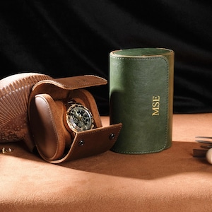 May include: A brown leather watch travel case with a watch inside, next to a green leather watch case. The watch case is open, revealing a gold watch with a black face. The green case has the initials "MSE" in gold.