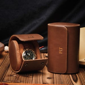 Op de afbeelding: Bruin leren horlogereisetui met een gouden horloge erin. De etui is open en onthult het horloge met een zwarte wijzerplaat. Een cilindrische horlogekist staat ernaast. De initialen "JM" staan op de etui.