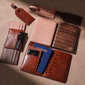 Puede incluir: Una variedad de accesorios de viaje de cuero en varios tonos de marrón y rosa. Los artículos incluyen carteras, fundas para pasaportes y etiquetas de equipaje con textura de piel de cocodrilo. Algunas carteras tienen ranuras para tarjetas y un pasaporte.
