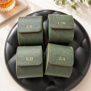 Puede incluir: Cuatro estuches de reloj de cuero verde con letras doradas, cada uno con las iniciales: JIR, CIW, SIB y JIA. Los estuches están colocados sobre una bandeja negra texturizada. Un vaso de líquido ámbar es visible en el fondo.