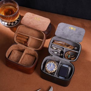 May include: Two open leather travel cases, one brown and one black, each with a soft interior. The brown case has the initials "S B" in gold on the lid. The black case holds a gold watch and a smartwatch.