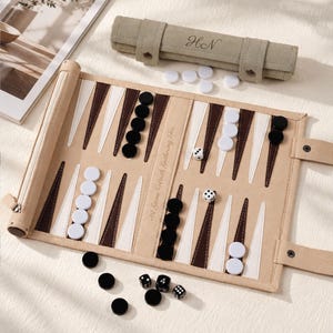 May include: A roll-up backgammon board in tan fabric with brown and white triangular points. The board features black and white circular checkers, two white dice, and three black dice. A matching tan fabric storage roll with the initials HN is included.