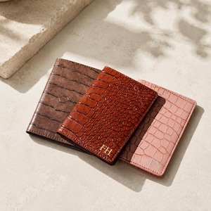 May include: Three rectangular passport holders with a crocodile-embossed texture. The left holder is dark brown, the center is reddish-brown with gold-colored initials FH, and the right is light pink with silver-colored initials TN.