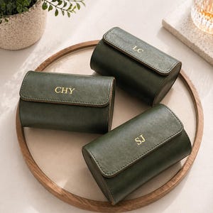 以下が含まれることがあります： 金色のイニシャル「LC」、「CHY」、および「SJ」が付いた、3つの濃い緑色の革製時計ケース。木製のトレイに配置されています。背景には植物とグラスがあります。これらのケースは時計を収納するように設計されています。