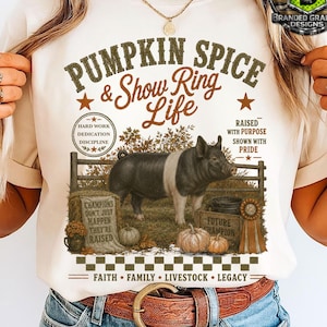 May include: A cream-colored t-shirt featuring a graphic of a black and white pig in a pen. The text reads PUMPKIN SPICE & Show Ring Life, HARD WORK DEDICATION DISCIPLINE, RAISED WITH PURPOSE SHOWN WITH PRIDE, CHAMPIONS DON'T JUST HAPPEN THEY'RE RAISED, FUTURE CHAMPION, FAITH FAMILY LIVESTOCK LEGACY.