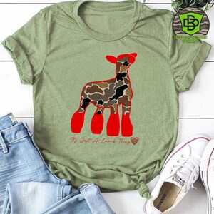 May include: Olive green t-shirt featuring a red and camouflage lamb graphic. The text "It's Just A Lamb Thing" is printed below the graphic, along with a heart symbol. The shirt is paired with blue jeans and white sneakers.