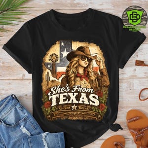 Peut inclure: T-shirt noir avec un motif d'une femme portant un chapeau de cow-boy et des lunettes de soleil, avec le texte "She's From TEXAS". Le motif comprend le drapeau am&eacute;ricain, un moulin &agrave; vent et des roses.