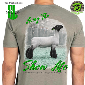 Può includere: Maglietta grigia con una grafica di una pecora con zampe e testa nere. Il design include il testo "Living The Show Life" in verde, con testo aggiuntivo in basso. La maglietta ha un logo tascabile gratuito.