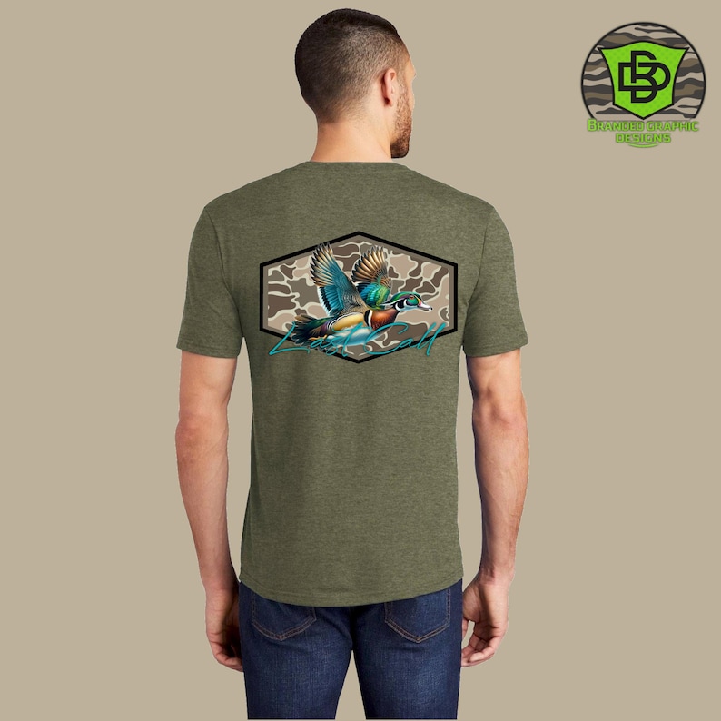 May include: Olive green t-shirt featuring a colorful illustration of a flying duck within a hexagonal frame with a camouflage background. The shirt has a crew neck and short sleeves. The back of the shirt displays the graphic design. The brand logo is in the upper right corner.