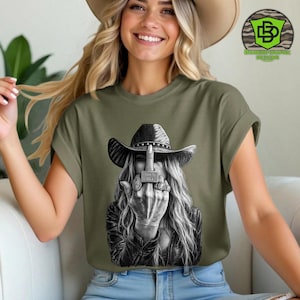 May include: Olive green t-shirt featuring a black and white graphic of a woman wearing a cowboy hat and leather jacket, flipping the middle finger. The graphic includes the text "Truth Hurts". The shirt has short sleeves and a relaxed fit.