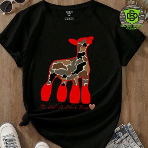May include: Black t-shirt featuring a graphic of a lamb with a camouflage pattern and red accents. The text "It's Just A Lamb Thing" is below the lamb graphic, with a heart symbol. The shirt has a crew neck and short sleeves.