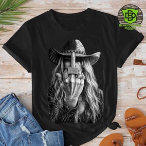 May include: Black t-shirt featuring a black and white graphic of a person wearing a cowboy hat and leather jacket, flipping the middle finger. The person is wearing multiple rings, one of which reads "F*** Off".