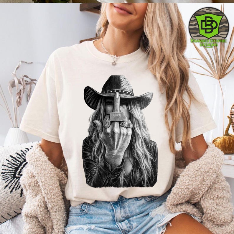 May include: Cream-colored t-shirt featuring a black and white graphic of a woman wearing a cowboy hat and leather jacket, flipping the middle finger. The graphic includes rings and the text "F*** It".