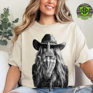 May include: Beige t-shirt featuring a black and white graphic of a person wearing a cowboy hat and leather jacket, giving the middle finger. The image includes the text "Fuck Off" on the middle finger and a logo in the upper right corner.