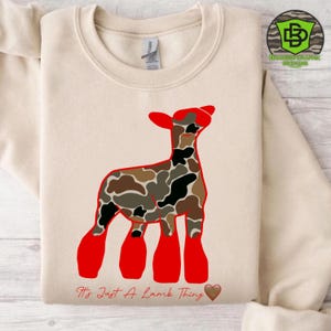 May include: A cream-colored sweatshirt featuring a red and camouflage lamb graphic. The text "It's Just A Lamb Thing" is printed below the lamb, with a brown heart. The sweatshirt has a crew neck and long sleeves.