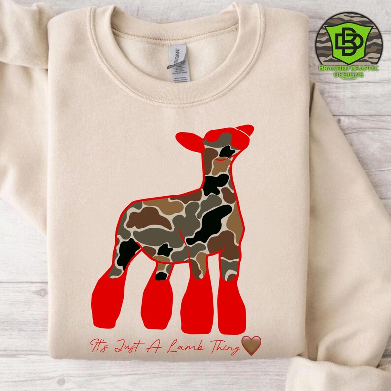 May include: A cream-colored sweatshirt featuring a red and camouflage lamb graphic. The text "It's Just A Lamb Thing" is printed below the lamb, with a brown heart. The sweatshirt has a crew neck and long sleeves.