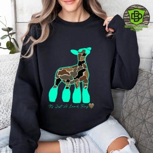 May include: Black sweatshirt featuring a cartoon lamb design with a camouflage pattern and teal accents. The text "It's Just A Lamb Thing" is below the lamb. A heart graphic is also included.