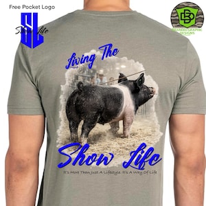 Può includere: Una maglietta grigia con un disegno di maiale in bianco e nero e le parole "Living The Show Life" in blu. Il testo "It's More Than Just A Lifestyle. It's A Way Of Life" è sotto. Un logo è nell'angolo in alto a destra.