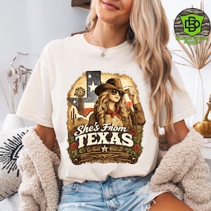 Peut inclure: T-shirt cr&egrave;me avec un motif d'une femme portant un chapeau de cow-boy et des lunettes de soleil, avec le texte "She's From TEXAS". Le motif comprend le drapeau du Texas, un moulin &agrave; vent et des roses.