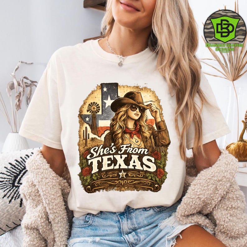 Peut inclure: T-shirt cr&egrave;me avec un motif d'une femme portant un chapeau de cow-boy et des lunettes de soleil, avec le texte "She's From TEXAS". Le motif comprend le drapeau du Texas, un moulin &agrave; vent et des roses.
