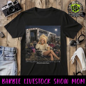 Puede incluir: Una camiseta negra con un estampado gr&aacute;fico de una mu&ntilde;eca de pelo rubio sentada en una silla plegable, sosteniendo una bebida, frente a un remolque de ganado. El texto dice: "Literally just give me a minute to find my sanity #Stocktrailerchaos".