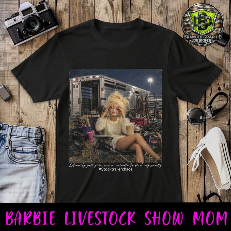 Puede incluir: Una camiseta negra con un estampado gr&aacute;fico de una mu&ntilde;eca de pelo rubio sentada en una silla plegable, sosteniendo una bebida, frente a un remolque de ganado. El texto dice: "Literally just give me a minute to find my sanity #Stocktrailerchaos".
