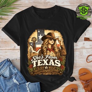 Peut inclure: T-shirt noir avec un graphisme repr&eacute;sentant une femme portant un chapeau de cowboy et des lunettes de soleil, avec le texte "She's From Texas". Le motif comprend le drapeau du Texas, des roses et un paysage d&eacute;sertique.