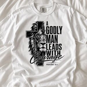 May include: A white short-sleeve t-shirt featuring a black graphic print of a lion's face partially overlaid by a cross. The text reads A GODLY MAN LEADS WITH Courage and PROVERBS 28:1 in a bold, distressed black font centered on the chest.
