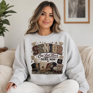 May include: A light gray crewneck sweatshirt featuring a graphic print with the text SENIOR One Last March BAND MOM and 2027. The design includes illustrations of a marching band hat, sheet music, a trumpet, and a drum on a football field.