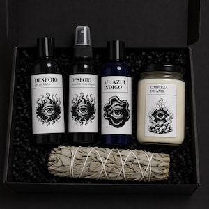 Puede incluir: Una caja de regalo negra que contiene tres botellas negras con etiquetas blancas, un frasco de vidrio con una vela blanca y un atado de salvia seca atado con hilo blanco. Las etiquetas muestran ilustraciones de ojos y el texto Despojo y Limpieza.
