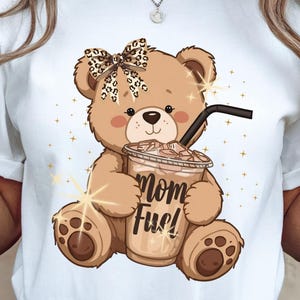 May include: White t-shirt featuring a cartoon teddy bear holding a coffee drink with the text "Mom Fuel." The bear has a leopard print bow and is surrounded by gold stars.