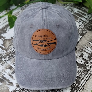 May include: Gray baseball cap with a curved brim and adjustable strap. A round, brown leather patch on the front features the text "If empathy is a sin, SIN BOLDLY" in white lettering, with a graphic of two hands shaking.