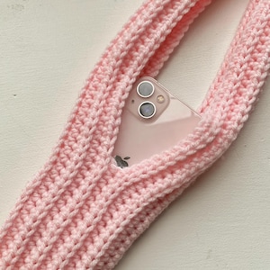Könnte beinhalten: Ein hellrosa gehäkelter Telefonhalter mit einer durchsichtigen Handyhülle im Inneren. Der Halter besteht aus dickem, strukturiertem Garn und hat ein Riemendesign. Das Telefon hat eine Dual-Kamera-Konfiguration.