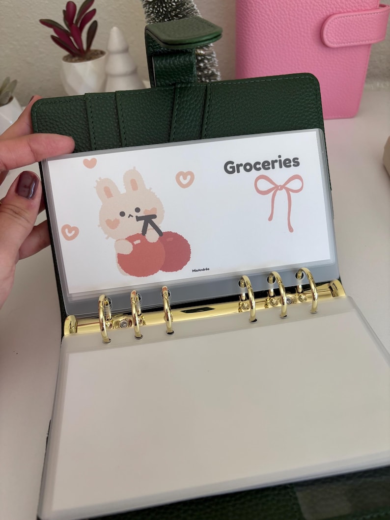 May include: A dark green binder with gold rings, open to reveal a clear pocket labeled "Groceries." The pocket features a cartoon bunny with cherries and hearts. A pink planner is visible in the background.