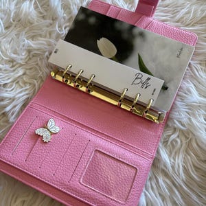 May include: Pink leather planner with gold-colored rings and a butterfly charm. The planner is open, revealing card slots and a clear ID window. Inside are inserts with the word "Bills" and a photo of a white tulip.