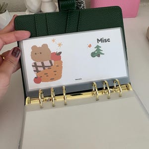 May include: A green leather binder with gold-colored rings, holding a clear plastic sleeve. The sleeve features a cartoon bear in a basket with apples, the word "Misc", and a small Christmas tree illustration.