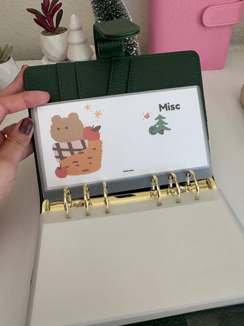 May include: A green leather binder with gold-colored rings, holding a clear plastic sleeve. The sleeve features a cartoon bear in a basket with apples, the word "Misc", and a small Christmas tree illustration.