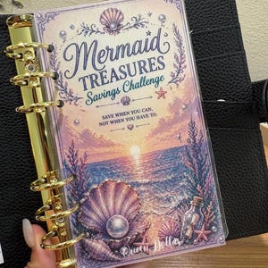 May include: A planner insert with the words "Mermaid Treasures Savings Challenge" on the cover. The design features a sunset over the ocean, seashells, and a pearl. The planner is held open with gold rings.
