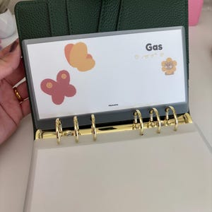 May include: A dark green binder with gold-colored rings, holding a clear plastic sleeve. The sleeve displays the word "Gas" and butterfly illustrations. The binder is open, revealing blank pages.