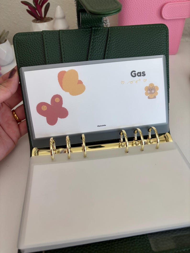 May include: A dark green binder with gold-colored rings, holding a clear plastic sleeve. The sleeve displays the word "Gas" and butterfly illustrations. The binder is open, revealing blank pages.